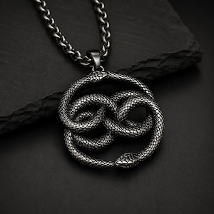Puede incluir: Un collar de color gris plomo oscuro con un colgante circular en forma de serpiente enroscada en un símbolo de infinito. La serpiente presenta un patrón de escamas detallado y texturizado en todo el cuerpo, uniendo cabeza y cola.