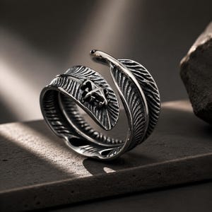 May include: A silver-toned metal ring shaped like a wrap-around feather with detailed etched lines and a small, dark, cross-like symbol embossed on the surface. The band is open at the top, showcasing the textured, curved design of the feather motif.