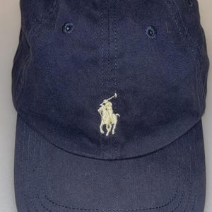 May include: A navy blue baseball cap featuring a white embroidered logo of a polo player on a horse centered on the front panel. The cap has a curved brim, a button at the top, and stitched eyelets for ventilation on the crown panels.