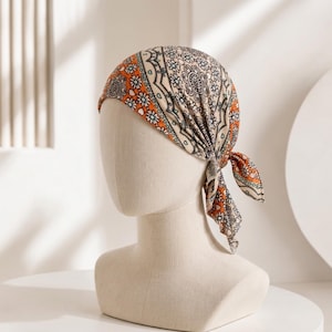 May include: A headscarf featuring a vibrant orange and cream paisley and floral pattern with black outlines. The fabric is tied at the back with two small, pointed ends hanging down, covering the top and sides of a beige mannequin head form.