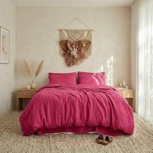May include: A vibrant magenta linen duvet cover set on a bed with two matching pillows. Above the bed hangs a large, triangular macrame wall hanging featuring dried palm leaves and floral elements. Two wooden nightstands flank the bed.