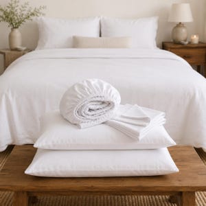 May include: A set of white bedding including a fitted sheet, two pillowcases, and a flat sheet. The fitted sheet is folded into a circular shape, while the flat sheet is neatly stacked on top of two rectangular pillows on a wooden bench.