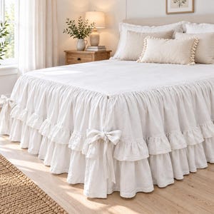 May include: A white bed skirt featuring a double-layered ruffled design with decorative fabric bows at the corners. The bed skirt is made of a textured fabric and drapes over the sides of the bed frame, creating a layered appearance.