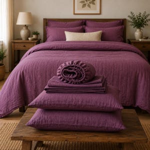 May include: A deep plum-colored bedding set featuring a textured duvet cover, two matching pillowcases, a folded flat sheet, and a fitted sheet. The bedding is made of a soft, crinkled fabric and is displayed on a wooden surface in front of a bed.