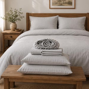Peut inclure: Un ensemble de literie gris clair composé d'un drap-housse, d'un drap plat et de deux taies d'oreiller empilés sur un banc en bois. L'ensemble présente une finition en tissu texturé, le drap-housse montrant des bords élastiqués au sommet.