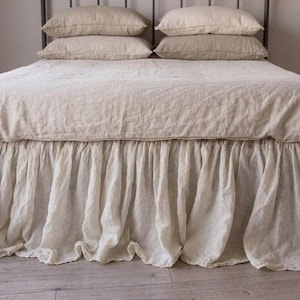 May include: A beige bed ensemble featuring a duvet cover and four matching pillows. The bed is adorned with a long, gathered bed skirt that reaches the floor, characterized by its soft, textured fabric and voluminous, flowing pleated design.