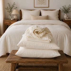 May include: A set of cream-coloured bedding including a fitted sheet, a flat sheet, and two pillowcases stacked on a rustic wooden bench. The bedding features a textured fabric finish and is displayed in front of a made bed with matching linens.