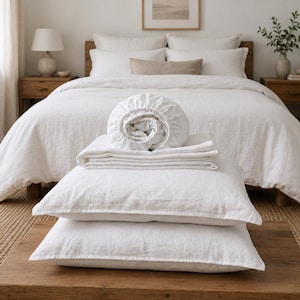 May include: A set of white linen bedding including two rectangular pillowcases, a folded flat sheet, and a fitted sheet with elastic edges, all stacked neatly on a wooden surface in front of a made bed with matching white linens.