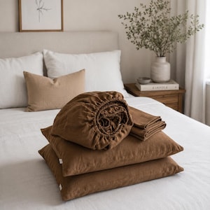 May include: A set of solid tobacco brown bedding including two rectangular pillowcases, a folded flat sheet, and a fitted sheet with elasticized edges, all featuring a visible woven texture and small white fabric tags on the side.
