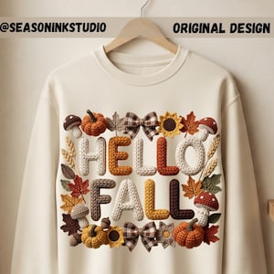 以下が含まれることがあります： クリーム色のクルーネックのスウェットシャツで、ニットのような質感の「HELLO FALL」という文字のグラフィックプリントが施されています。デザインの周囲には、カボチャ、キノコ、ヒマワリ、秋の葉、チェック柄のリボンのイラストが描かれています。