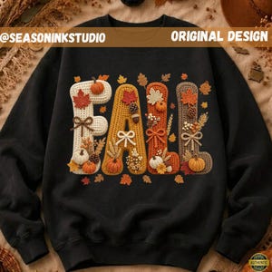 May include: A black long-sleeve sweatshirt featuring the word FALL in large, textured, crochet-style letters. Each letter is decorated with small pumpkins, pinecones, wheat stalks, bows, and autumn leaves in shades of cream, orange, and brown.