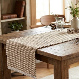 May include: A rectangular table runner featuring a textured, woven pattern in a neutral beige and off-white colour palette. The runner is laid flat across a wooden table, showcasing a consistent, dense knit construction with a finished edge border.