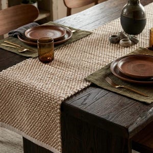 May include: A beige and white woven table runner with a textured pattern, placed on a dark wooden table. The setting includes olive green fabric placemats, brown ceramic plates, gold-toned cutlery, amber glassware, and a two-toned ceramic vase.