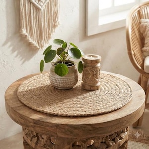 May include: A round woven jute placemat sits on a carved wooden side table. On the placemat, there is a small green Pilea peperomioides plant in a speckled ceramic pot and a cylindrical wooden candle holder with intricate carved patterns.