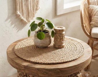 May include: A round woven jute placemat sits on a carved wooden side table. On the placemat, there is a small green Pilea peperomioides plant in a speckled ceramic pot and a cylindrical wooden candle holder with intricate carved patterns.
