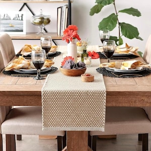 May include: A wooden dining table set for a meal with a beige textured table runner, black woven placemats, white plates, yellow and white plaid napkins, wine glasses, a wooden bowl of blue corn chips, and a vase with red gerbera daisies.