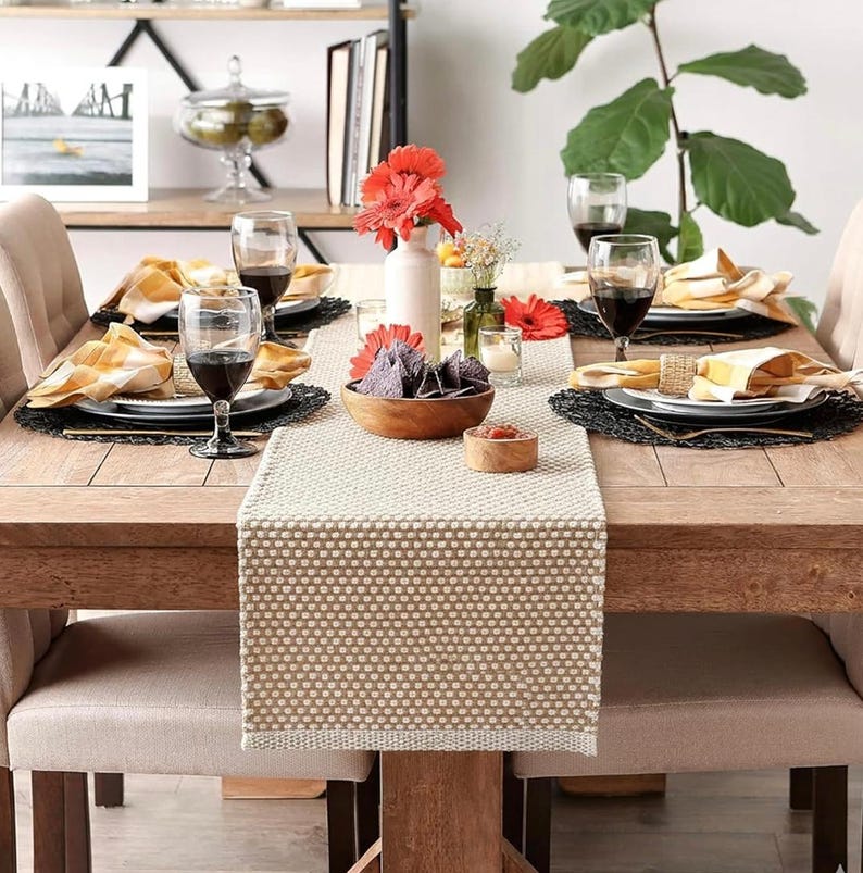 May include: A wooden dining table set for a meal with a beige textured table runner, black woven placemats, white plates, yellow and white plaid napkins, wine glasses, a wooden bowl of blue corn chips, and a vase with red gerbera daisies.