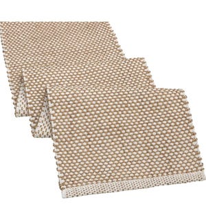 May include: A rectangular table runner featuring a woven texture with a repeating pattern of tan and off-white threads. The fabric displays a dense, grid-like weave structure with a finished edge border in a lighter off-white tone on the ends.