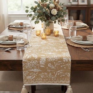 May include: A mustard yellow table runner featuring a white floral pattern and cream-colored tassels at both ends. The runner is placed on a wooden dining table set with white plates, woven placemats, clear glassware, and wooden napkin rings.
