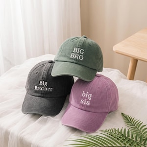 Puede incluir: Tres gorras de béisbol en verde salvia, gris carbón y morado polvoriento. La gorra verde salvia presenta el texto bordado en blanco BIG BRO. La gorra gris carbón muestra el texto bordado en blanco Big Brother. La gorra morada tiene big sis.