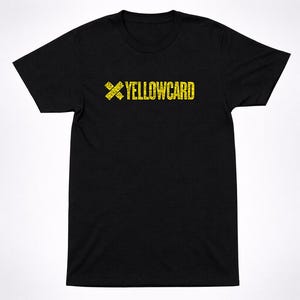 May include: A black short-sleeved t-shirt featuring a yellow graphic on the chest. The graphic consists of a stylized X symbol followed by the text YELLOWCARD, both rendered in a distressed, textured yellow print against the black fabric.