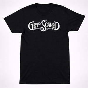 Può includere: Una t-shirt nera a maniche corte con la scritta GET SCARED stampata in un carattere bianco, stilizzato e decorativo sul petto. La maglietta presenta un girocollo e una vestibilità standard, con la scritta bianca centrata sul tessuto nero.