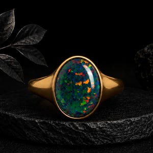May include: A gold ring featuring a large, oval-shaped dark opal gemstone. The stone displays a vibrant play of color with flecks of red, orange, and green against a deep black background. The band is smooth, polished gold with a simple, solid design.