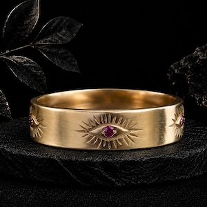 May include: A gold band ring featuring a repeating engraved eye motif with a small, round, vibrant pink gemstone set in the center of each eye. The metal surface has a smooth, matte finish, and the design encircles the entire outer circumference.