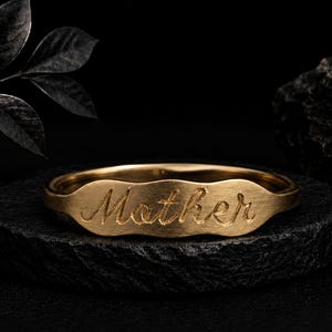 Könnte beinhalten: Ein goldfarbener Ring mit einer flachen, gewellten Mittelplatte, in die das Wort "Mother" in Schreibschrift eingraviert ist. Der Ring hat eine matte Oberfläche und liegt auf einem dunklen, strukturierten runden Sockel neben einem Blatt.