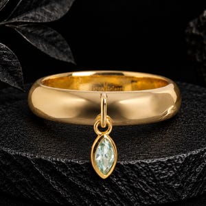 Puede incluir: Un anillo de banda de oro pulido con un dije colgante individual. El dije es una piedra preciosa de color verde claro con corte marquesa engastada en un marco de oro, suspendido de una pequeña argolla de oro unida a la superficie lisa y redondeada.