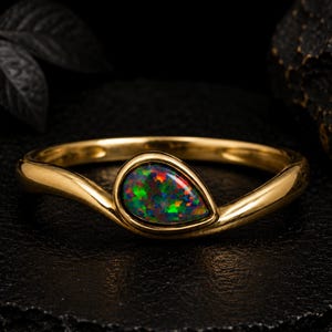 Pode incluir: Um anel de ouro com uma única pedra preciosa de opala negra em forma de pera. A opala exibe um jogo de cores vibrante com reflexos vermelhos, verdes e laranja. A banda é feita de ouro amarelo polido e curva-se em torno da pedra em gota.