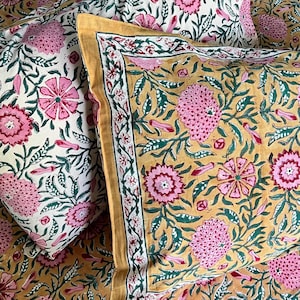May include: Two square pillow shams featuring a floral block print pattern. One sham has a cream background, while the other has a mustard yellow background. Both display pink flowers and green leaves with a decorative border around the edges.