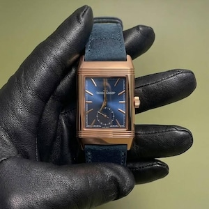 Könnte beinhalten: Eine rechteckige Armbanduhr mit einem Gehäuse aus Roségold und einem tiefblauen Zifferblatt mit Sonnenschliff. Die Uhr verfügt über ein kleines Sekunden-Hilfszifferblatt unten, goldfarbene Indizes und den Schriftzug Jaeger-LeCoultre. Sie hat ein blaues Wildlederarmband.