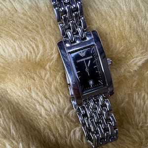 May include: A silver-toned Emporio Armani wristwatch featuring a rectangular case and a black dial with silver hands and markers. The watch includes a date window at the six o'clock position and a multi-link metal bracelet with a polished finish.