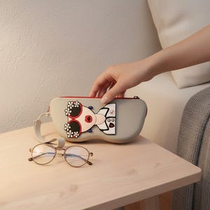 May include: A light grey hard-shell glasses case with a zip closure and a wrist strap. The case features a graphic of a face wearing red-rimmed sunglasses decorated with white flowers and red lips. A pair of thin-rimmed metal glasses sits in front.