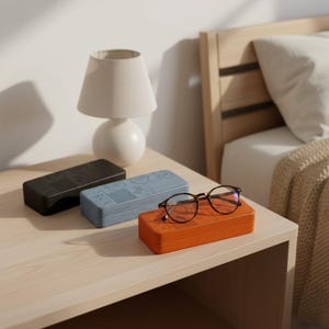 Puede incluir: Tres cajas de almacenamiento rectangulares en negro, azul y naranja, cada una con una cuadrícula de patrones geométricos en relieve en la tapa. Un par de gafas de montura oscura y fina descansa sobre la caja naranja, sobre una superficie de madera clara.