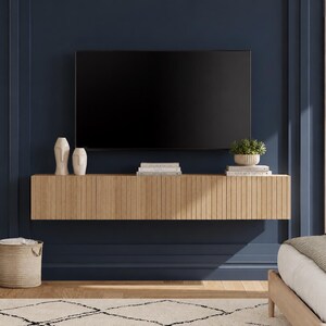 May include: A wall-mounted floating media console featuring a light wood finish with vertical slats. A flat-screen television is mounted above the console, which holds two abstract white vases, a small potted green plant, and stacked white books.