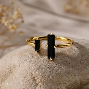May include: A gold-toned open ring featuring two black baguette-cut stones. The larger stone is set vertically on one end, while the smaller stone is set horizontally on the opposite end of the polished metal band, creating a distinct gap.