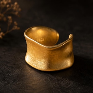 May include: A wide, open-ended gold cuff ring with a textured, brushed finish. The band features an irregular, undulating wave-like shape along the top and bottom edges, creating a fluid, organic form with a matte, metallic yellow appearance.