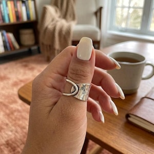May include: A silver ring featuring a wide, hammered-texture band that wraps around the finger, connected by a smooth, curved silver wire element that creates an open, layered design on the side of the finger, showcasing a metallic finish.