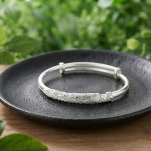 May include: A silver-toned bangle bracelet featuring an intricate, embossed feather or leaf pattern along the front section. The bracelet has a smooth, rounded band design with a visible overlapping mechanism that allows for size adjustment.