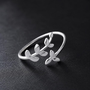 May include: A silver-toned open ring featuring a delicate branch design with multiple small, smooth leaves extending along the band. The metal has a polished, reflective finish, and the ring is shaped to wrap partially around the finger.