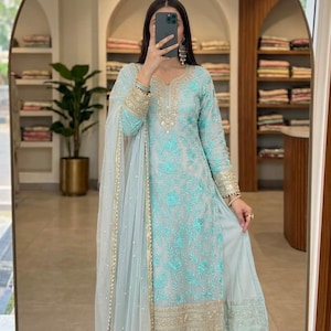 May include: A light blue long-sleeved tunic featuring intricate turquoise floral embroidery and gold metallic detailing along the neckline, cuffs, and hem. The outfit includes matching wide-leg trousers and a sheer, lightweight dupatta with gold trim.