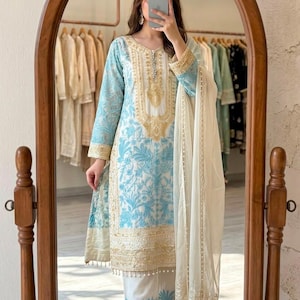Könnte beinhalten: Ein dreiteiliges Set bestehend aus einer langärmeligen Tunika, einer gerade geschnittenen Hose und einem transparenten Dupatta. Die weiße Tunika ist mit hellblauen Blumenstickereien und Goldfäden verziert, passend zum Muster der Hose.