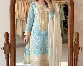Puede incluir: Un conjunto de tres piezas que consta de una túnica de manga larga, pantalones de corte recto y un dupatta transparente. La túnica blanca presenta bordados florales en azul claro y detalles en hilo dorado, a juego con el pantalón.