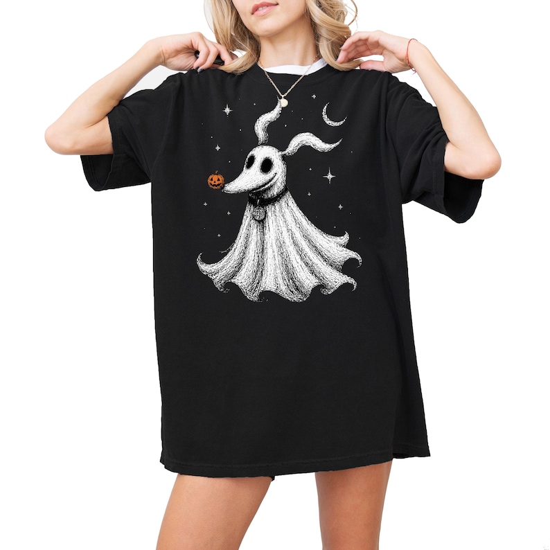 Op de afbeelding: Een zwart T-shirt met korte mouwen en een witte grafische illustratie van een spookhond met lange oren, een donkere halsband met een penning en een kleine oranje pompoen op de neus, omringd door witte sterren en een halve maan op de borst.