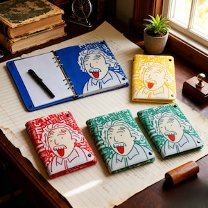 May include: A collection of five ring-bound notebooks featuring a cartoon illustration of Albert Einstein with his tongue out. The covers come in blue, yellow, red, and green, all patterned with the E=mc² equation. One blue notebook is open with a black pen.