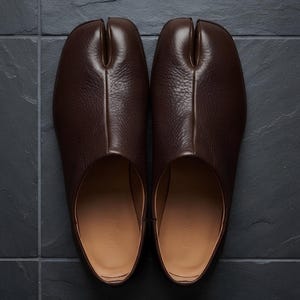 May include: A pair of dark brown leather shoes featuring a distinct split-toe design. The shoes have a textured exterior surface, a smooth tan interior lining, and a low-profile silhouette. The design separates the big toe from the other toes.