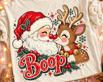 Könnte beinhalten: Ein cremefarbenes T-Shirt mit einer Grafik von einem Weihnachtsmann und einem Rentier, die sich an den Nasen berühren. Darunter steht das Wort Boop in großen roten Buchstaben. Das Design zeigt Stechpalmen, rote Beeren und grüne Schneeflocken.