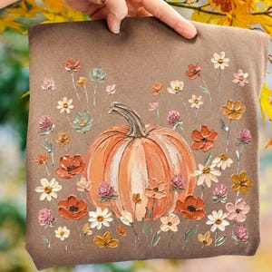 May include: A brown t-shirt featuring a central orange pumpkin illustration surrounded by a variety of painted-style flowers in shades of pink, orange, yellow, and white. The design uses thick, textured brushstrokes to create a dimensional effect.